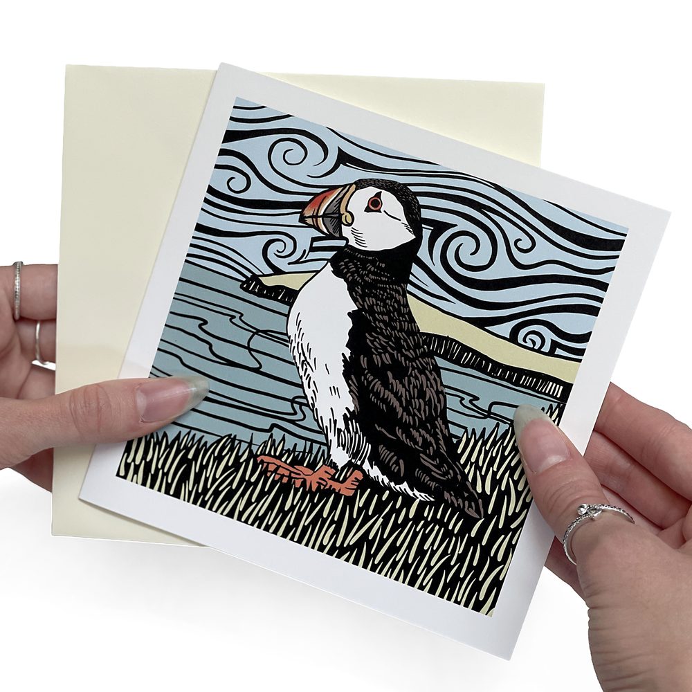 A square linocut-style puffin greeting card with a cream envelope