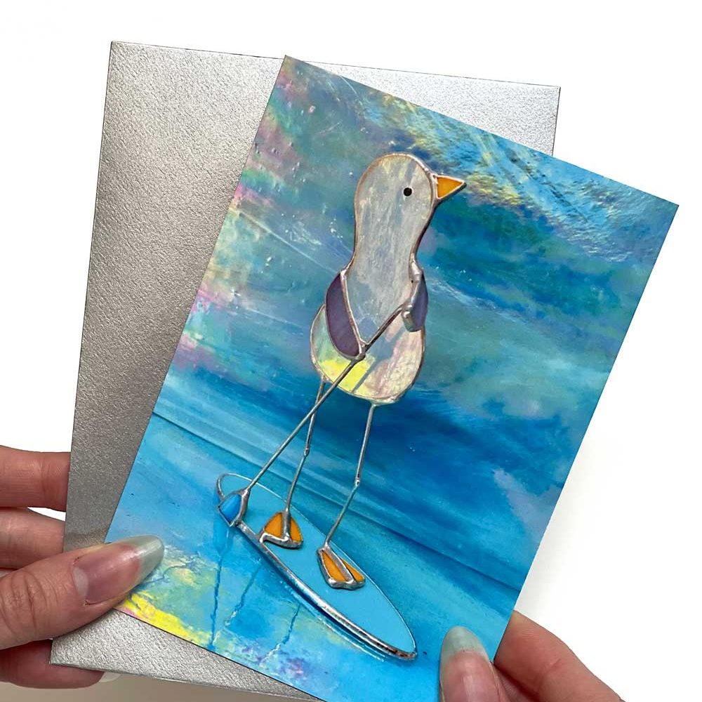 A portrait greeting card of a stained-glass seagull on a surfboard, with a silver envelope