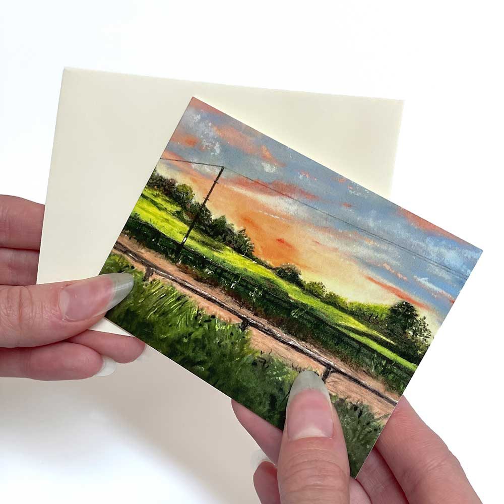 A landscape greeting card of a sunset over fields and railway, with a cream envelope