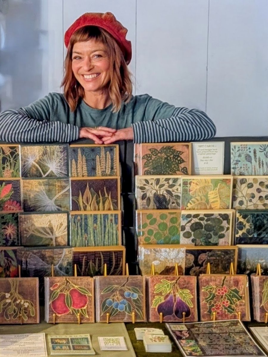 Jacqui Caley-Hawker of Alcemi Art smiling behind her market display of botanical art cards
